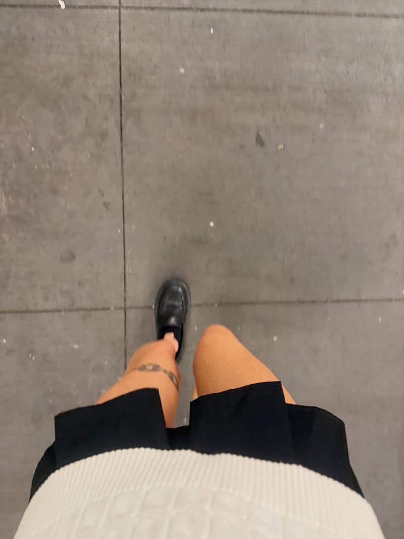 Looking down at a pleated skirt and black loafers on a city sidewalk