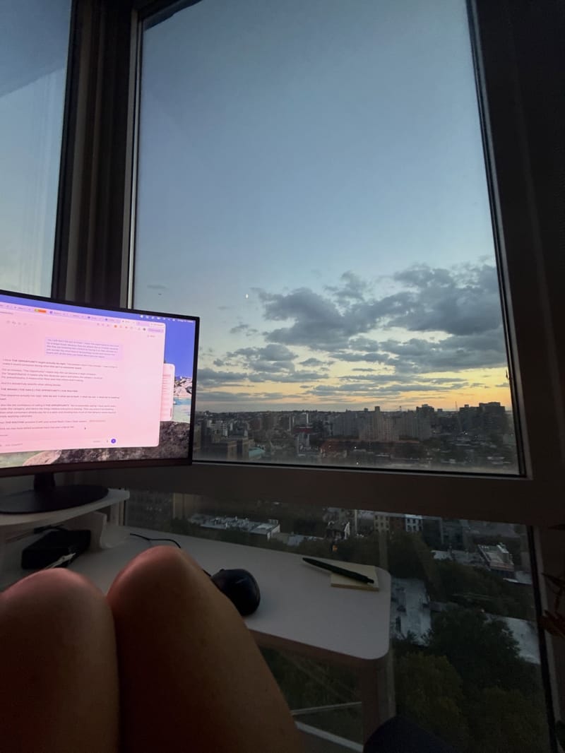 A desk with a monitor beside a window at sunset over the city